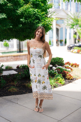 A smiling woman posing outdoors on a sidewalk wearing the Capri Citrus & Palms Midi Dress. The white A-line dress features an all-over whimsical postcard motif with illustrations of lemons, palm trees, crabs, and vintage stamps. It is detailed with dainty spaghetti straps and a wide, tan geometric lattice border framing both the square neckline and the bottom hem. She styles the look with white thong sandals and a minimal gold bar pendant necklace against a lush garden backdrop.