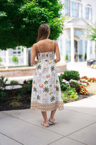 Back view of the Capri Citrus & Palms Midi Dress showcasing its flattering, secure construction. The dress features a wide, flexible smocked panel across the upper back that transitions into a fluid A-line shift silhouette. The whimsical travel graphics and the rich tan-and-white geometric border hemline drape beautifully to a sophisticated midi length, paired with white flat slides.