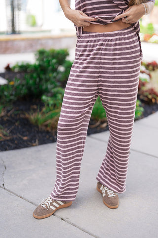 A detailed front close-up view focusing on the waist and leg construction of the Wildberry Boardwalk Pants. The image highlights the flexible, pull-on elastic waistband that sits comfortably on the waist and the relaxed drape of the wide legs. The soft knit fabric displays a distinctive heathered texture across the alternating mauve and soft pink stripes, styled with classic athletic sneakers.