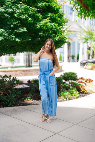 Woman smiling and posing outdoors on a concrete sidewalk wearing the Boardwalk Bound Strapless Romper. The lightweight, mineral-washed blue jumpsuit features a secure strapless neckline, an elasticized drawstring waist, and an architectural wide-leg silhouette with prominent exposed seam detailing. Styled for a casual streetwear aesthetic with flat white thong sandals with gold medallions and a gold chain necklace.