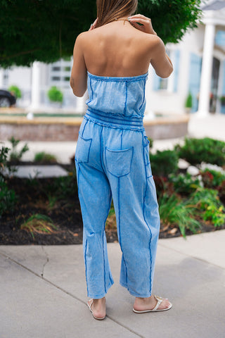 Back view of the Boardwalk Bound Strapless Romper showcasing the relaxed fit and structural barrel leg drape from behind. The mineral-washed blue fabric features an elasticized top band and a gathered waist that flows into a roomy silhouette, detailed with two symmetrical rear patch pockets and finishing at a contemporary cropped ankle length.
