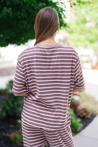 Back view of the Wildberry Boardwalk Tee illustrating its unlined, fluid construction from behind. The continuous mauve and dust pink stripe pattern runs horizontally across the shoulders and relaxed straight hemline. The model stands slightly turned on the walkway, showing how the oversized shape pairs effortlessly with the matching knit lounge bottoms.