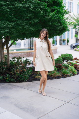 Woman smiling and posing outdoors on a concrete sidewalk wearing the Upper East Side Pocket Dress. The sleeveless, elegant cream-colored mini dress features a structured pointed collar and a gentle, relaxed A-line silhouette. Styled for a preppy, Hamptons-inspired look with flat white leather thong sandals and delicate gold bracelets against a lush green tree backdrop.