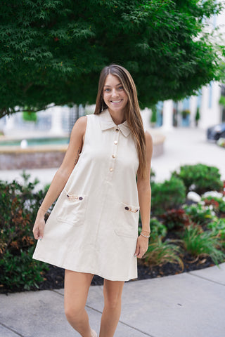 A mid-shot front view of the Upper East Side Pocket Dress highlighting its relaxed yet sophisticated shift silhouette. The model smiles while lightly holding the hem of the crisp cream woven dress, showcasing the partial button-front placket with polished gold-tone buttons and symmetrical front patch pockets.