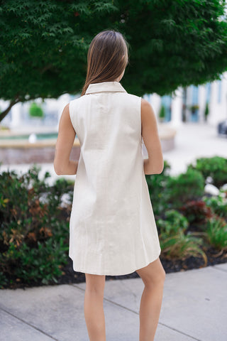 Back view of the Upper East Side Pocket Dress showcasing the clean, uninterrupted shift drape of the lightweight woven cream fabric. This view emphasizes the timeless sleeveless profile and a neatly folded structured pointed collar from behind as the model poses outdoors next to garden landscaping.