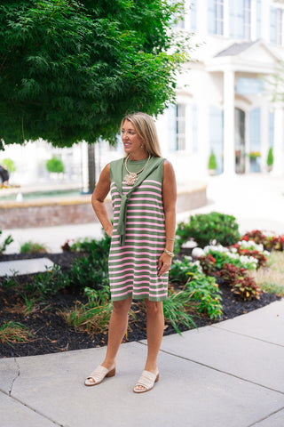 Woman posing outdoors on a sidewalk wearing the Tulip & Twine Striped Dress, a sleeveless sweater knit mini dress featuring alternating pink and sage green horizontal stripes. The preppy shift-style dress showcases a built-in, attached solid sage green knit scarf tie detail draped smoothly over her shoulders and loosely knotted at the front. She stands with one hand on her hip, completing the country-club look with cream-colored low block-heeled slide mules and a gold statement starburst necklace.