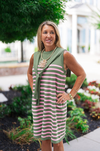 A three-quarter portrait view highlighting the unique neckline structure and premium texture of the Tulip & Twine Striped Dress. The shot emphasizes the clean round crew neckline and the high-quality, lightweight sweater knit fabrication that skims comfortably over the torso. The attached sage green shoulder-tie detail creates a sophisticated layered illusion, beautifully complemented by layered gold bracelets and a striking textured pendant necklace.