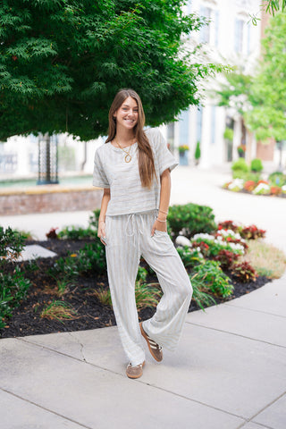 A full-length front view of a woman smiling and posing outdoors in the Ocean Breeze Lounge Set. The matching sky blue and cream knit set features an effortless, boxy short-sleeve top and loose, fluid wide-leg lounge pants. She is styled for a chic travel look with brown and white retro sneakers and a layered gold link statement necklace, standing on a sidewalk with a lush green backdrop.