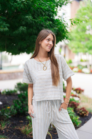 A mid-length front view of the Ocean Breeze Lounge Set top, showcasing its wide round neckline, relaxed dropped short sleeves, and unique front patch pocket. The lightweight, multi-directional textured knit highlights the subtle cream and sky blue stripes, contrasted by a diagonal stripe pattern on the chest pocket. The look is accessorized with a prominent gold hoop pendant necklace.
