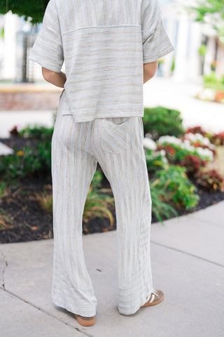 A full-length back view of the Ocean Breeze Lounge Set, showcasing the comfortable drape of both the boxy short-sleeve top and wide-leg trousers. The back perspective highlights a single rear patch pocket on the pants detailed with a playful diagonal stripe layout. The lightweight, non-sheer sky blue and cream fabrication is paired with casual low-profile sneakers.