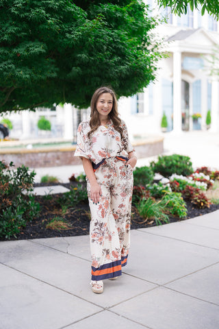 Woman smiling and posing outdoors on a sidewalk wearing the Venice Vineyard Blouse, styled as a high-end, coordinated resort set with the matching wide-leg trousers. The lightweight woven cream blouse features a relaxed boxy silhouette adorned with a vintage botanical floral print. She completes the look with cream platform wedge sandals and a delicate gold necklace against a lush garden backdrop.