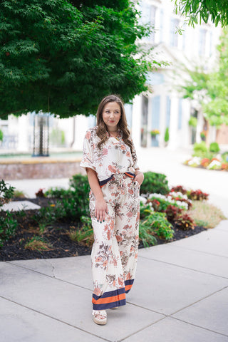 Woman posing outdoors in the Venice Vineyard Pants, a pair of billowy, wide-leg resort trousers featuring a cream base with an intricate vintage botanical floral print in terracotta, dusty rose, and olive tones. The trousers are styled as a coordinated luxury set with the matching short-sleeve button-up blouse. She stands on a sidewalk next to garden landscaping, completing the look with cream-colored platform wedge sandals and gold jewelry.