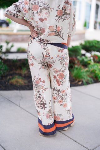 Back view focusing on the Venice Vineyard Pants to showcase the pattern placement and fluid drape of the lightweight woven fabric. The shot highlights how the vintage botanical floral print covers the pants and coordinating blouse, with the bold navy and terracotta orange border framing the bottom hemline right above the woven platform sandals.
