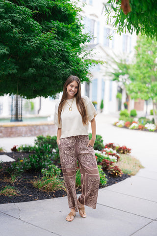 A smiling woman posing outdoors on a concrete path wearing the Java Jungle Wide-Leg Pants. The breezy, lightweight trousers showcase an artistic neutral animal print in coffee brown, warm tan, and soft cream tones. Styled for an upscale, safari-inspired resort look, she pairs the billowy wide-leg pants with a short-sleeve cream knit top, an oval gold link necklace, and flat tan leather thong sandals.