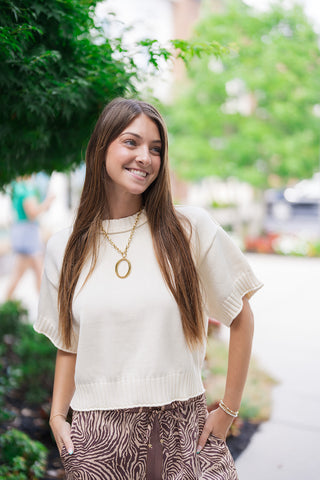 A medium portrait view of the Coastline Short Sleeve Sweater highlighting its premium, ultra-soft knit texture and detailed construction. The image captures the classic round crew neckline, distinct dropped shoulders, and chunky ribbed trim framing the neckline, short sleeve cuffs, and straight waist hemline. The model looks off-camera with her hands in her pockets, showing off a laid-back, sophisticated aesthetic.