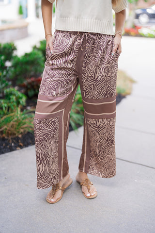 A detailed front close-up of the Java Jungle Wide-Leg Pants highlighting the unique, high-end scarf-print placement. The abstract zebra-print motif is segmented by a solid, mocha-brown contrast border running vertically down the center seams and horizontally across the legs. This shot emphasizes the flexible elasticated waistband with its matching adjustable drawstring and the model's hands tucked inside the discreet side pockets.