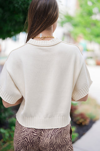 Back view of the Coastline Short Sleeve Sweater showcasing the clean, unlined drape of the lightweight ivory-cream knit fabric. The shot emphasizes the comfortable slouchy shape across the shoulders and the structured look of the thick ribbed knit borders at the neck, sleeves, and hemline as the sweater sits smoothly over patterned trousers.