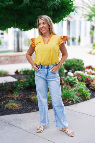 A woman posing outdoors on a sidewalk wearing the Saffron Sunsets Flutter Top. The mixed-media top features a vibrant marigold yellow slub knit body with contrasting terracotta orange woven ruffle sleeves. Styled with her hands in her pockets, she pairs the relaxed-fit blouse with high-rise, light-wash denim straight-leg jeans and cream-colored flat slide sandals.