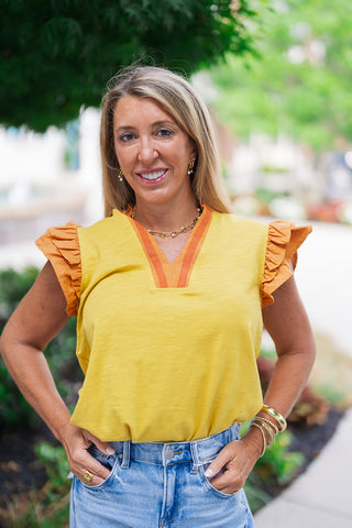 A portrait view of the Saffron Sunsets Flutter Top highlighting its unique textures and neckline. The close-up showcases the organic slub knit texture of the yellow bodice and the sharp split V-neckline framed by a structured, terracotta striped trim placket. The model is accessorized with a gold link necklace, small hoop earrings, and stacked gold bracelets.