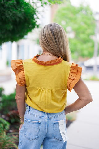 Back view of the Saffron Sunsets Flutter Top showcasing the clean fit and double-layered shoulder volume from behind. The marigold bodice features a gathered yoke detail across the upper back that transitions into a relaxed drape, while a contrasting terracotta striped trim outlines the back of the ruffled collar. The shirt is neatly tucked into high-waisted denim.