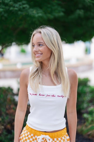A mid-length front view showcasing the modern silhouette of the Stadium Social Tank. The sleeveless white tank top highlights a flattering square neckline and vertical ribbed knit texture. The front displays the phrase "I'm just here for the tailgate" in detailed, textured crimson script embroidery. The tank is styled untucked over vibrant orange and white checkered athletic shorts with a white drawstring.