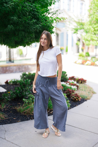 A woman posing outdoors on a concrete path wearing the Stormy Seas Wide-Leg Pants in a slate-blue hue. The trousers feature a thick smocked waistband with an adjustable drawstring and a relaxed, billowy wide-leg silhouette. Styled for a casual coastal vibe, she pairs the textured resort pants with a simple tucked white t-shirt and flat white leather thong sandals in front of a landscaped garden backdrop.