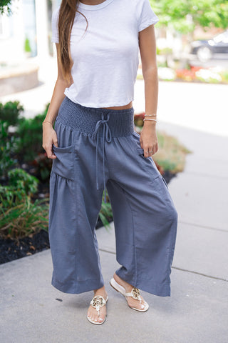 A detailed front close-up shot focusing on the unique construction of the Stormy Seas Wide-Leg Pants. The view highlights the wide, elasticized smocked waist, the functional front drawstring feature, and the lightweight, beachy texture of the slate-blue fabric. Symmetrical, oversized dropped pouch pockets are visible along the sides of the billowy silhouette, styled with gold medallion thong sandals.