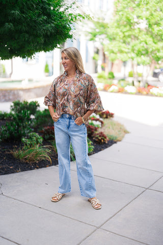 A smiling woman posing outdoors on a concrete sidewalk wearing the Canyon Spice Botanical Top. The relaxed-fit bohemian blouse features an allover botanical leaf print in a warm "Red Clay Mix" palette. Styled for a chic daytime look, she stands with her hands in the pockets of her high-waisted, light-wash wide-leg denim jeans, finished with cream-colored flat slide sandals.
