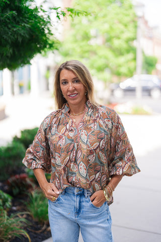 A full-length front view of the Canyon Spice Botanical Top showcasing its relaxed drape and fluid hemline. The blouse is casually half-tucked into light-blue denim jeans, highlighting the structural balance created by the voluminous 3/4 length statement puff sleeves. The model smiles at the camera against a landscaped garden background.