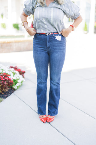A mid-body front view focusing on the tailored fit of the high-rise, dark wash flare jeans. The denim is styled with a half-tucked black and white micro-check blouse, secured at the waist with a thin red belt featuring gold hardware. The model's hands rest at her waistline, highlighting the smooth front fit of the jeans and a Good American brand tag tucked into the front left pocket. Glossy red pointed-toe heels anchor the bottom of the wide-leg hem.