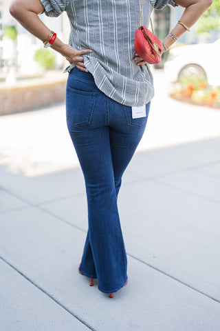 Back view of the Good American dark wash jeans, showcasing the flattering contoured seat, classic back pocket placement, and elegantly flared hem. A white Good American brand tag is attached to the back right pocket. The checkered puff-sleeve blouse is left untucked in the back, displaying its relaxed, breezy drape. The model rests her hands on her hips, with the red quilted crossbody bag hanging smoothly by her side above the red pointed-toe heels.
