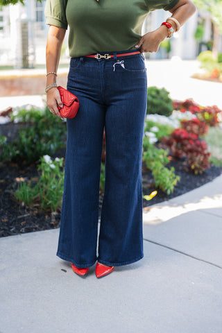 Close-up front view focusing on the tailored fit of dark wash, high-rise wide-leg jeans. The denim is styled with a tucked-in olive green knit top, secured at the waist with a thin red belt featuring gold hardware. The model holds a small red quilted handbag, perfectly coordinating with the glossy red pointed-toe pumps peeking out from the wide denim hem.