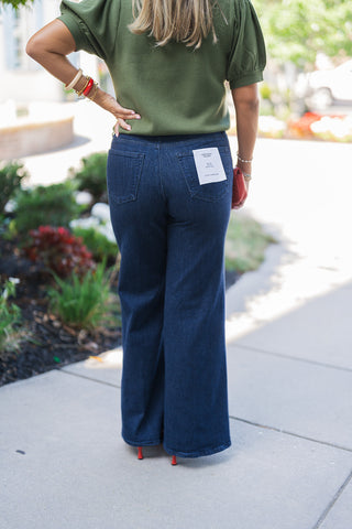 Back view of the Good American Good Waist Palazzo jeans in a dark wash, highlighting the smooth, high-rise fit and classic back pocket placement. The wide-leg denim drapes cleanly down to a pair of red pointed-toe heels. The model wears a tucked-in olive green short-sleeve sweater, with a hand resting casually on her hip.