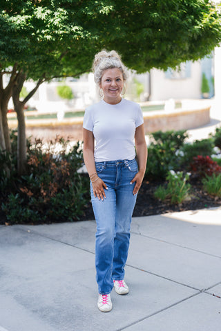 Woman smiling outdoors in a casual, everyday outfit featuring a crisp white short-sleeve t-shirt and medium wash straight-leg jeans. The relaxed look is accessorized with large gold hoop earrings, layered bracelets, and white distressed sneakers with vibrant neon pink laces. Styled in front of a green tree and paved courtyard, the image conveys a classic, effortless aesthetic.