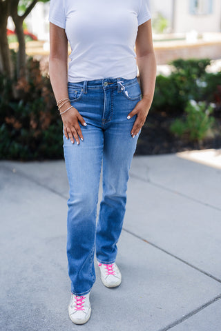 Close-up front view focusing on the flattering fit of the medium wash straight-leg jeans. The classic denim is styled with a simple white t-shirt and features subtle fading along the thighs with standard front pocket styling. The model rests her hands on her upper legs, highlighting her white manicure and layered bracelets, while white sneakers with bright pink laces anchor the bottom of the frame.