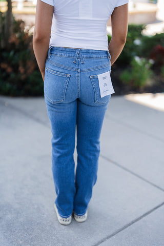 Back view of the Good American Good Legs Straight jeans in a medium wash, highlighting the contoured back yoke and classic back pocket placement. A visible white tag on the right pocket confirms the Good American brand and straight-leg style. The straight-leg denim drapes cleanly down to a pair of white sneakers featuring animal print heel details, paired seamlessly with the white t-shirt.