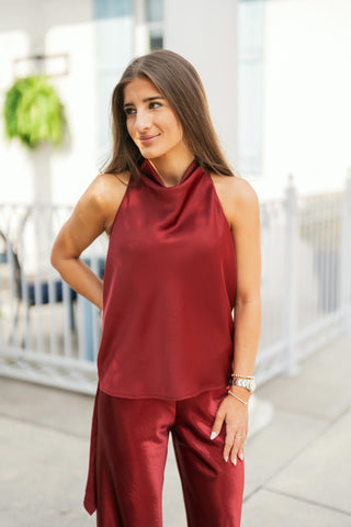 Woman posing outdoors wearing a deep burgundy satin sleeveless halter top featuring a high draped neckline with soft cowl pleating. The lustrous top is styled as part of a monochromatic set paired with matching high-waisted burgundy satin wide-leg pants. She accessorizes with stacked silver and gold beaded bracelets, a watch, and delicate rings against a bright, modern architectural background.