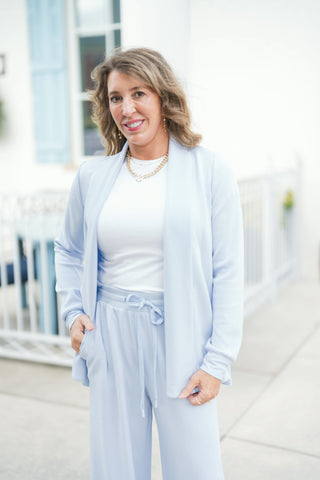 Woman posing outdoors wearing a light blue soft-knit open-front cardigan featuring a lapel-style drape collar, drop shoulders, and long ribbed-cuff sleeves. The cozy cardigan layer is worn over a fitted white crew-neck tee and paired with matching light blue wide-leg drawstring lounge pants, styled with a gold link necklace and drop earrings against a bright building exterior.