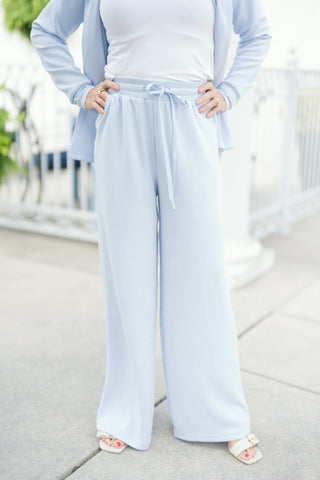 Lower-body shot of a woman modeling light blue soft-knit wide-leg lounge pants featuring a high-waisted elastic waistband with an adjustable drawstring tie and side slanted pockets. The relaxed pants drape smoothly to a wide hemline, paired with a fitted white crew-neck tee, a matching light blue open-front cardigan, and off-white buckled slide sandals on an outdoor paved walkway.