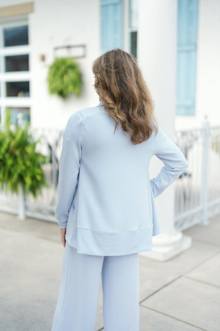Back view showing the rear silhouette of the light blue cardigan set, highlighting a wide ribbed bottom hem band, smooth back knit panel, and relaxed sleeve cut. The model stands with one hand on her hip, showcasing the longline hem that falls gracefully over the matching light blue wide-leg trousers.