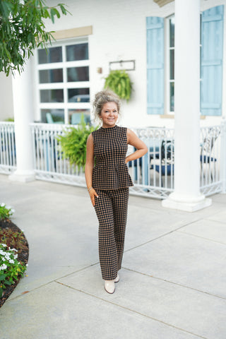 Woman standing outdoors wearing a matching two-piece knit set in a tan and black polka dot pattern. The ensemble features a sleeveless, mock-neck peplum top with a flared hem paired with high-waisted, wide-leg knit trousers. Styled with cream patent leather ankle boots, gold hoop earrings, and a curly updo in front of a white building background.