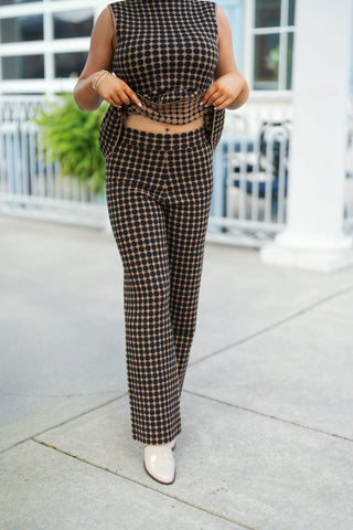 Close-up shot showing the high-waisted fit and straight, wide-leg cut of the tan and black polka dot knit pants. The model gently lifts the peplum hem of the matching sleeveless top to highlight the smooth elastic waistband and soft stretch knit fabric. Paired with cream pointed-toe boots.