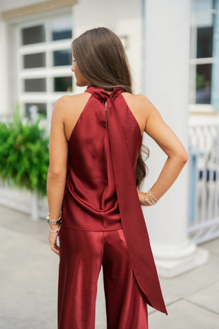 Back view of the burgundy satin halter top highlighting a high tie-neck detail with long, dramatic sash tails extending down the back. The backless sleeveless silhouette seamlessly pairs with matching burgundy satin trousers, showing the fluid drape and glossy finish of the fabric styled with layered beaded bracelets and a silver watch.