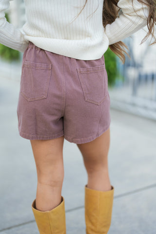 A back close-up view highlighting the functional short silhouette of the dusty mauve skort, featuring an comfortable elastic waistband and two classic back patch pockets. Shows the back hem of the cream ribbed sweater and pairs seamlessly with knee-high tan leather boots.