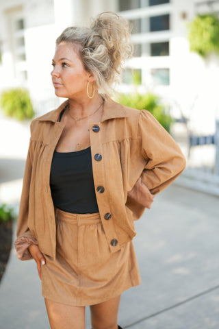 Three-quarter view of the tan faux suede jacket showcasing its rich, matte texture, structured spread collar, and prominent dark contrast buttons. The shot highlights the gentle gathering at the chest yoke and puffed shoulder seams, styled layered over a fitted black tank top with delicate gold hoop earrings.