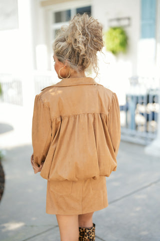 Back view of the tan faux suede jacket highlighting the romantic gathered back yoke that creates a subtle, voluminous drape. Features slightly slouched shoulders, long sleeves with turned-up cuffs, and a clean lower hemline, worn over a matching tan suede mini skirt with a curly blonde updo.