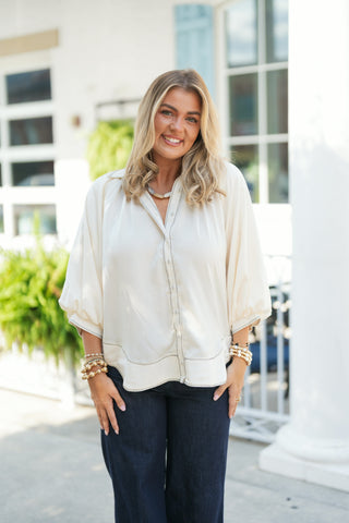 Woman smiling outdoors wearing a cream silky satin button-down blouse featuring dark contrast whipstitch stitching along the placket, collar, hemline, and sleeve cuffs. The top is designed with voluminous three-quarter length balloon sleeves, an open V-neckline collar, and an easy, relaxed curved hem. Styled with dark-wash jeans, stackable gold and beaded bracelets, and a stone necklace in front of a white building background.