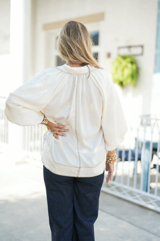Back view of the cream satin blouse highlighting the intricate contrast cross-stitching along the gathered back neck seam, vertical center spine, and rounded hemline trim. Demonstrates the airy volume of the raglan balloon sleeves, worn with hand on hip paired with dark-wash denim jeans.