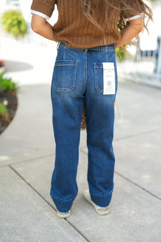 Back view detailing the rear construction of the medium-dark wash straight-leg jeans, showcasing angular back yoke stitching, two flat patch pockets, and a neat raw hem resting at the top of white sneakers. The model stands with hands tucked near her waist, wearing a brown cable-knit sweater with white sleeve trim.