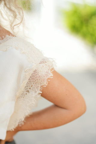 A detailed close-up shot highlighting the intricate off-white eyelash lace trim along the shoulder and cap sleeve of the cream satin top. The fine, floral-patterned lace edge contrasts softly against smooth skin, emphasizing the delicate craftsmanship and airy texture of the garment's trim.